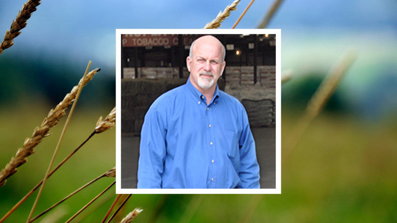 A man in a blue shirt stands inside a warehouse. The background shows wheat stalks in a blurred field, suggesting an agricultural setting.