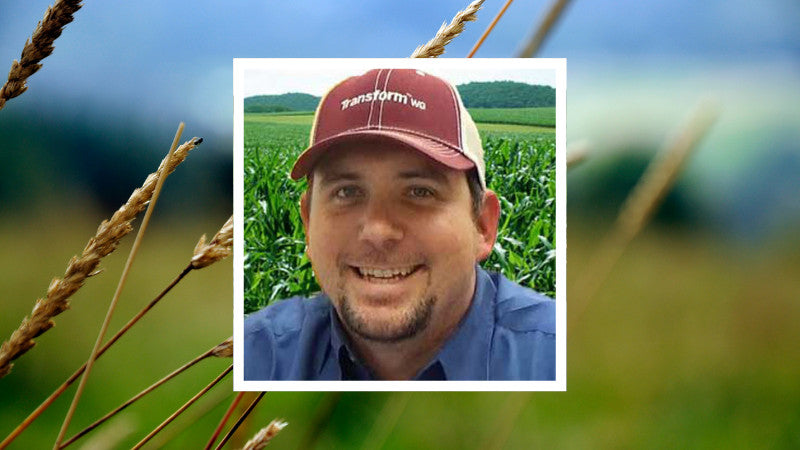 Smiling man wearing a red cap and blue shirt, framed in white. Behind him, a lush green cornfield and wheat stalks convey a rural farming vibe.