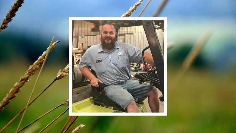 A smiling man wearing a blue work shirt and denim shorts sits on a forklift in a workshop, framed by a background of blurred wheat fields.